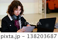 Focused teenage girl with headphones around neck studies on laptop at large table, holding spiral notebook and working on school assignment, concept online education and digital learning for teens. 138203687