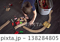 Boy Tidying Wooden Train Set On Dark Floor, Reaching Into Basket To Store Tracks And Accessories, Overhead 138284805