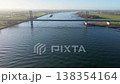 Aerial view of a modern cable stayed bridge crossing a wide river at sunrise. Green meadows, river groynes, and distant wind turbines under a hazy blue sky. 138354164