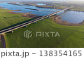 Aerial panoramic view of a modern cable stayed bridge spanning a wide river in a rural European landscape. Long cargo barges navigate the water surrounded by lush green fields and winding roads. 138354165