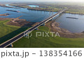 Aerial panoramic view of a modern cable stayed bridge spanning a wide river in a rural European landscape. Long cargo barges navigate the water surrounded by lush green fields and winding roads. 138354166