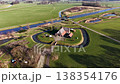 High angle drone view of a straight Dutch canal cutting through vibrant green pastures, featuring a traditional farmhouse, a small water control gate, and a parallel cycling path. 138354176