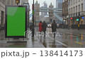 A digital billboard with a green screen stands on a wet London street, with Tower Bridge visible. 138414173