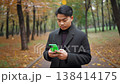 A young Asian man walks through a park in autumn, engrossed in his smartphone. 138414175