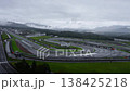 Aerial daytime view of Fuji Speedway near Mount Fuji. White cars run wet laps, spray rises on rain slick asphalt. Potenza and Bridgestone branding appears along track. 138425218