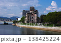 Hiroshima Peace Memorial stands by the Motoyasu River as tourists stroll with parasols. A bridge, office buildings, and hills frame the dome under cumulus clouds. 138425224