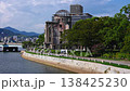 Daytime view of the A Bomb Dome on the Motoyasu River, visitors stroll with umbrellas, blue bridge and city buildings behind, park trees and low mountains in Hiroshima, Japan. 138425230