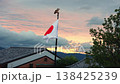 A white Hinomaru flag with deer finial flutters above gray kawara roofs and a leafy tree in Kyoto, with gabled buildings, a TV antenna, low mountains, and warm sunset light. 138425239