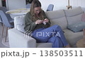 A woman sits on a sofa at home and knits with yarn, enjoying a calm and cozy moment while creating 138503511