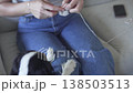 A woman sits on a sofa at home and knits with yarn while a small dog stays nearby, creating a cozy 138503513