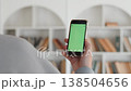 Close-up of a hand holding a smartphone with a mockup green screen, back view, with the background of white bookcases with many books on the bookshelves. 138504656