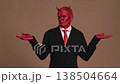 Businessman wearing devil horns and red makeup makes various hand gestures against a brown background. Expressions range from confusion to anger, showcasing themes of temptation and decision making 138504664