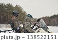 Riders accelerate snowmobile across snow, camouflage duo leaning forward, engine roar and snow spray, atv companion nearby, pine forest boundary, fast motion and freedom vibe during winter run 138522151