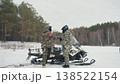 Two white men by snowmobile exchanging handshake and words, snowy open field near pine forest, full winter gear and helmets, one mounting vehicle while partner offers support and checks engine, 138522154