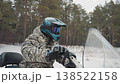 Rider on atv in snowy forest camouflage jacket and blue helmet with mirrored goggles gripping handlebars behind clear windshield scout checking trail pine trees cold winter light subtle breath visible 138522158