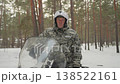 Snowmobile snowy pine forest rider stands beside vehicle in camouflage gear and helmet, engine idling, tracks cutting fresh snow, tall pines framing scene, soft snowfall and cold light convey rugged 138522161