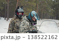 Two riders inspect atv before departure, camouflage coats and helmets with visors, snowy forest clearing, driver checks windshield and throttle, passenger reviews checklist, teamwork briefing, gear 138522162