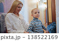 Mom fastening son sweater in hallway. Quiet tender scene of caring mother helping toddler into striped sweater while seated by doorway and mirror, gentle conversation and soft indoor light create cozy 138522169