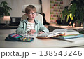 A schoolboy in glasses is thinking it over his homework at table in home setting. He is sitting in his bright room at white table, where his textbooks, copybook and an open pencil case filled with 138543246