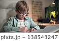 A child wearing glasses sits at a table in a home setting, and works on exercises in a notebook. He is sitting in bright room at a white table, where his textbooks and copybook . Close up shot 138543247