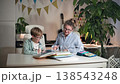 A mother and son are sitting in a bright room at a white table, upon which textbooks and the boy notebook are laid out, alongside an open pencil case filled with pencils and pens. The mother is 138543248