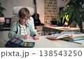 A little boy wearing glasses is doing his homework at a table in a home setting. He is sitting in his bright room at white table, where his textbooks, copybook and an open pencil case filled with 138543250