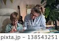 A mother and son are sitting at a table, she is explaining exercises to her son and helping him with his homework, she is correcting his mistakes. They a sitting in bright room at a white table 138543251