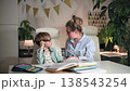 A mother and son are sitting at a table, she is explaining exercises to her son and helping him with his homework. They a sitting in bright room at a white table , where boys textbooks, copybook and 138543254