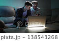 At home in a dark room, two friends are chatting. One of the guys is wearing glasses holding a mobile phone. They are sitting on a couch and watch video on smartphone with a laptop on a coffee table 138543268