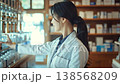 Professional asian pharmacist in a white lab coat working in a modern pharmacy. She carefully searches the shelves for a specific medication and then presents it, smiling at the camera 138568209