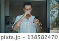 Modern lifestyle man standing at sliding glass door using smartphone and drinking juice during calm morning at home. Relaxed man checking messages on mobile phone while enjoying peaceful start of day. 138582470