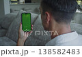 Man holding smartphone with green screen chroma key in vertical position, sitting on sofa at home. Male using mobile phone mockup with blank green display ready for app interface or video replacement 138582483