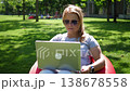 A young woman with a laptop is sitting in an easy chair in a modern park. The camera is stabilized, and the wind is blowing her hair. A freelance opens her laptop to take pictures of the park. 138678558