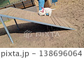 A West Highland White Terrier walks down a wooden ramp at an outdoor dog park. This is part of an agility training session for the dog. The park is designed for canine activities. 138696064