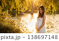 A woman stands serenely by the lake, her belly wrapped in a maternity dress, with the setting sun behind her. 138697148