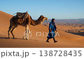 A Moroccan guide dressed in a blue robe gracefully walks alongside a camel across vibrant sand dunes. The warm hues of sunset illuminate the tranquil desert landscape, creating a serene atmosphere. 138728435