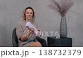 joyful woman, unwrap gift, cozy outfit. Joyful woman in cozy outfit sitting on chair, smiling warmly while unwrapping small rectangular gift box on table. 138732379