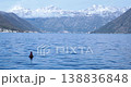 Snowcapped mountain peaks tower over Bay of Kotor Montenegro with red buoy floating in blue sea water. Distant coastal towns sit below foggy slopes on bright winter morning. 138836848