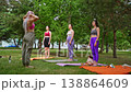 Women in yoga poses reaching toward floor in sunny park group class with green background. Yoga pose, forward fold, group, park, women, sunny. 138864609