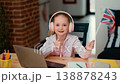 A girl with light skin sits at a table with a laptop. She wears headphones and smiles while holding a small British flag. She shows a thumbs up to express her happiness with online learning. 138878243