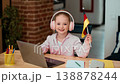 A girl with light skin is smiling while wearing headphones. She holds a small German flag in one hand. She is seated at a wooden desk with a laptop and stationery around her. 138878244