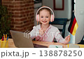 A girl sits at a table with a laptop, wearing headphones and smiling. She holds a small German flag in one hand and gives a thumbs-up, indicating success in her language learning. 138878250