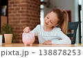 A young girl with light skin sits at a table, counting coins with a pink piggy bank nearby. A cup is placed next to her in a bright, cozy room with a brick wall. 138878253