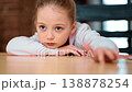 A young girl with light brown hair is resting her chin on her hands while sitting at a wooden table. She looks contemplative in a relaxed indoor environment with soft lighting. 138878254