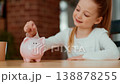 A girl with light skin smiles as she drops a coin into her pink piggy bank. She sits at a wooden table in her living room. This moment captures financial learning and saving. 138878255