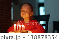 A young girl with light skin sits at a table, looking at a birthday cake with lit candles. The warm light creates a joyful atmosphere as she prepares to make a wish during a birthday celebration. 138878354
