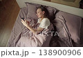 High angle view of a smiling young woman relaxing in bed and browsing her social media feed on a mobile phone after waking up in the morning, enjoying a peaceful start to her day 138900906