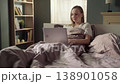 Relaxed young woman sitting comfortably in her bed under the duvet, smiling while working or browsing on her laptop computer early in the morning with soft sunlight coming through the window 138901058