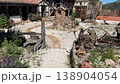 A rustic area with stone paths leading to a traditional building. There are plants, clay pots, and old tools for display. The sun shines on this outdoor location. 138904054