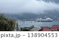 White Azamara cruise ship sails across calm bay waters in Montenegro. Foggy mountains tower over coastal town buildings during overcast day. Passenger ship moves slowly 138904553